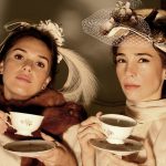 Two women in vintage attire enjoying a tea party, showcasing elegant fashion and hats.